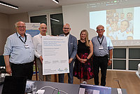 Fünf Personen stehen in einem Büro vor einem großen Plakat mit Text und lächeln in die Kamera