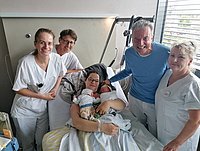 Eine Frau hält ein Neugeborenes in einem Krankenhausbett, umgeben von fünf Personen in weißen Kitteln. Die Personen stehen und sitzen nahe bei ihr.