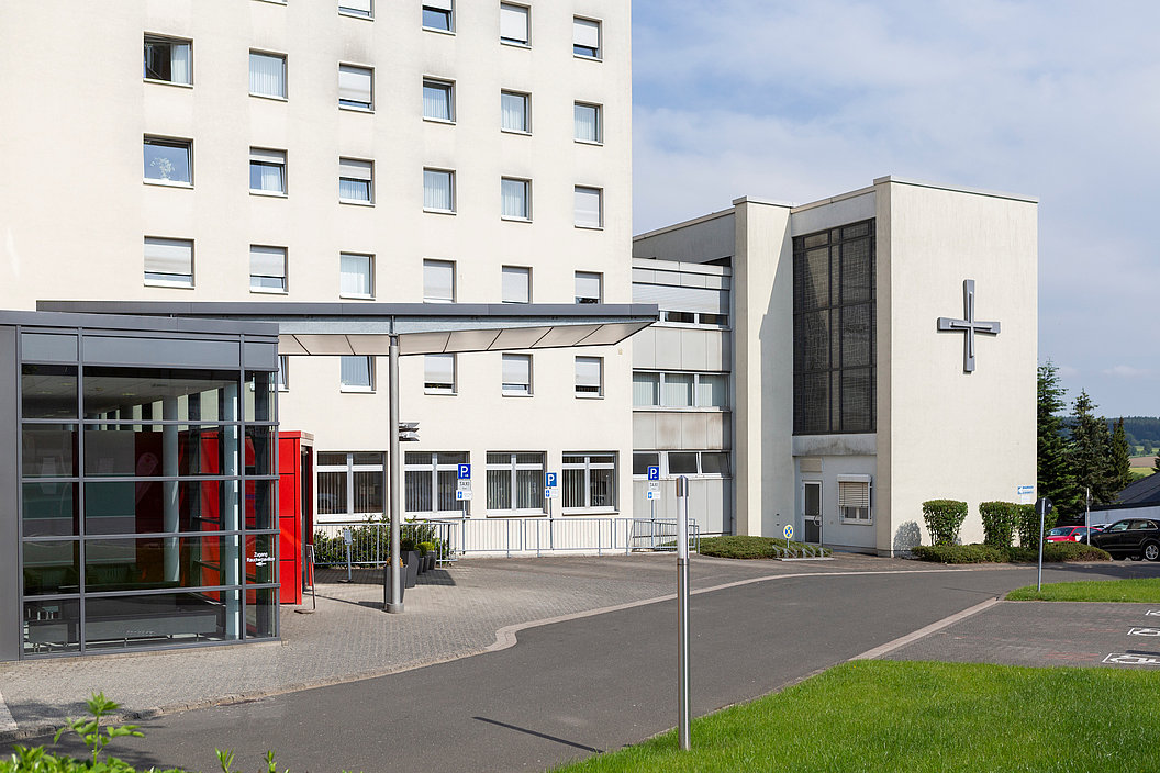 Außenansicht eines mehrstöckigen Krankenhausgebäudes mit Glasvorbau, rotem Briefkasten und großem Kreuz an der Fassade.