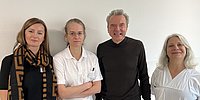 Vier Personen stehen nebeneinander vor einer hellen Wand, zwei Frauen links tragen helle Oberteile, ein Mann in der Mitte trägt ein dunkles Oberteil, eine Frau rechts trägt ein weißes Oberteil