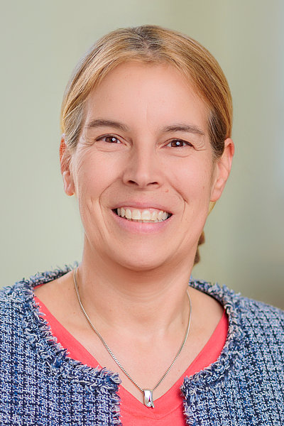 Frau mit blonden Haaren, blauem Strickpullover und rotem Oberteil, trägt eine silberne Halskette