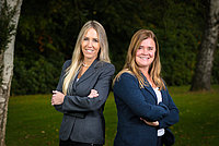 Zwei Frauen stehen nebeneinander im Freien, beide mit verschränkten Armen. Die Frau links trägt einen grauen Blazer, die Frau rechts einen blauen Blazer.