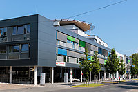Moderne, mehrstöckige Gebäude mit großen Fenstern und bunten Fassadenelementen. Vor dem Gebäude stehen Bäume und Parkplätze.