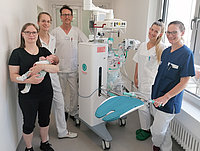 Sechs Personen in medizinischer Kleidung stehen in einem Raum. Eine Person hält ein Baby, während eine andere ein medizinisches Gerät mit einer blauen Liege präsentiert.