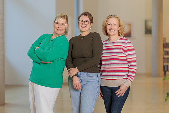 Drei lächelnde Frauen stehen nebeneinander in einem hellen Raum, die linke trägt eine grüne Bluse und weiße Hose, die mittlere eine schwarze Bluse und Jeans, die rechte einen rot-weiß gestreiften Pullover und dunkle Hose