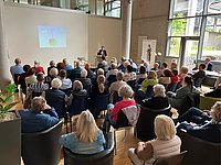 Publikum sitzt in einem modernen Raum und hört einem Vortragenden zu, der vor einer Leinwand steht