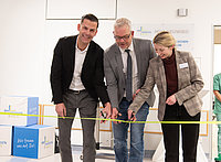Drei Personen in Businesskleidung schneiden gemeinsam ein gelbes Band durch, im Hintergrund stehen Kartons und eine Person in grüner Arbeitskleidung.