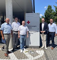 Fünf Männer in Hemden und Hosen stehen vor einem Schild mit rotem Kreuz und Text vor einem Gebäude unter blauem Himmel