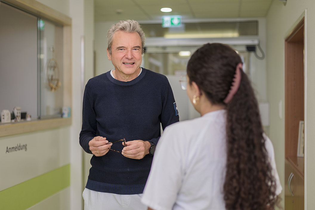 Ein Mann mit dunklem Pullover hält eine Brille in einem Krankenhausflur, vor ihm steht eine Frau mit langem, dunklem Haar und weißer Arbeitskleidung