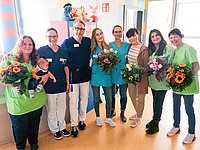 Gruppe von sieben Personen in Arbeitskleidung, die Blumensträuße halten. Eine Person hält ein Baby. Die Kleidung ist grün, blau und weiß.