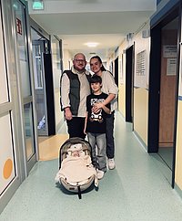 Drei Personen stehen in einem Flur, ein Kind hält einen Kinderwagen mit einer Decke darin