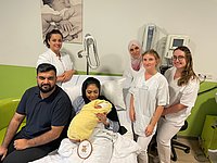 Gruppe von sechs Personen in einem Krankenhauszimmer, eine Frau hält ein in eine gelbe Decke gewickeltes Baby auf dem Schoß, umgeben von fünf weiteren Personen, von denen vier weiße Pflegekleidung tragen