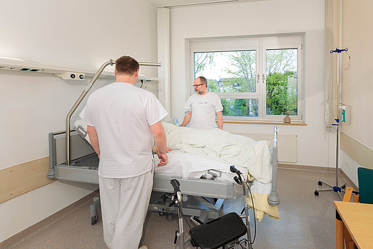 Zwei Männer in weißer Arbeitskleidung stehen an einem Krankenhausbett in einem hellen Patientenzimmer mit Fenster