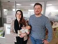 Junges Paar steht am Empfang des Kreißsaals, die Frau hält ein schlafendes Baby im Arm, beide blicken in die Kamera