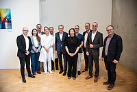 Gruppe von zehn Personen in formeller Kleidung, stehend in einer hellen Umgebung. Im Hintergrund ein farbenfrohes Kunstwerk an der Wand.