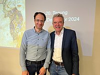 Dr. Korkolis und Dr. Berger stehen nebeneinander, beide in formeller Kleidung. Im Hintergrund ist ein Projektor mit dem Text '20. Tagung Dezember 2024' sichtbar.