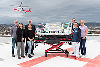 Eine Gruppe von sechs Personen steht um eine Tragbahre auf einem Hubschrauberlandeplatz. Im Hintergrund fliegt ein Rettungshubschrauber. Die Tragbahre ist mit medizinischen Geräten ausgestattet.
