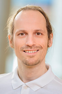 Frank Martin Kügler trägt ein weißes Polohemd und hat lange, blonde Haare, die zu einem Zopf gebunden sind. Der Hintergrund ist unscharf.