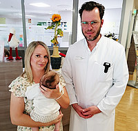 Eine Frau hält ein Baby in ihren Armen, während ein Mann in einem weißen Kittel neben ihr steht. Im Hintergrund sind Pflanzen und Dekorationen sichtbar.