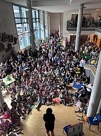 Große Menschenmenge in einem mehrstöckigen Innenraum mit Säulen und großen Fenstern, eine Person steht vorne und spricht zu den Sitzenden