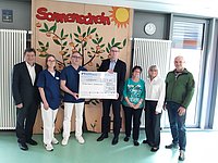 Gruppe von sieben Personen steht vor einer Wand mit einem Baum und der Aufschrift 'Sonnenschein', zwei Personen halten einen großen symbolischen Scheck