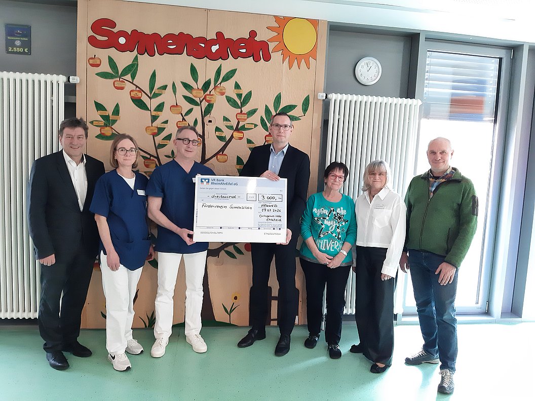 Gruppe von sieben Personen steht vor einer Wand mit einem Baum und der Aufschrift 'Sonnenschein', zwei Personen halten einen großen symbolischen Scheck
