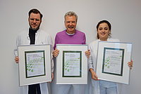 Drei Personen in weißen Laborkitteln halten gerahmte Zertifikate. Die Person in der Mitte trägt ein lila Oberteil. Die Zertifikate zeigen grüne Blätter und Text.