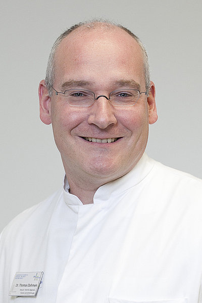 Dr. Dahmen, Oberarzt und Facharzt für Allgemein- und Gefäßchirurgie, trägt weißen Arztkittel, Hemd mit Krawatte und schwarze Brille