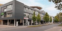 Moderne, mehrstöckige Gebäude mit großen Fenstern und einem begrünten Dach. Vor dem Gebäude stehen mehrere Bäume und Autos. Eine Straße verläuft entlang der Vorderseite.