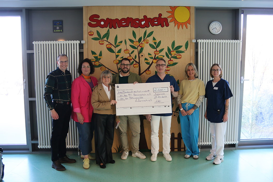 Gruppe von sieben Personen steht in einem Raum vor einer Wand mit einem großen bunten Wandbild mit der Aufschrift 'Sonnenschein' und hält einen großen symbolischen Scheck
