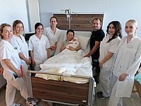Eine Gruppe von neun Personen in weißen Kitteln steht um ein Bett im Krankenhaus. Eine Person hält ein Baby, während die anderen lächeln und in Richtung der Kamera schauen.