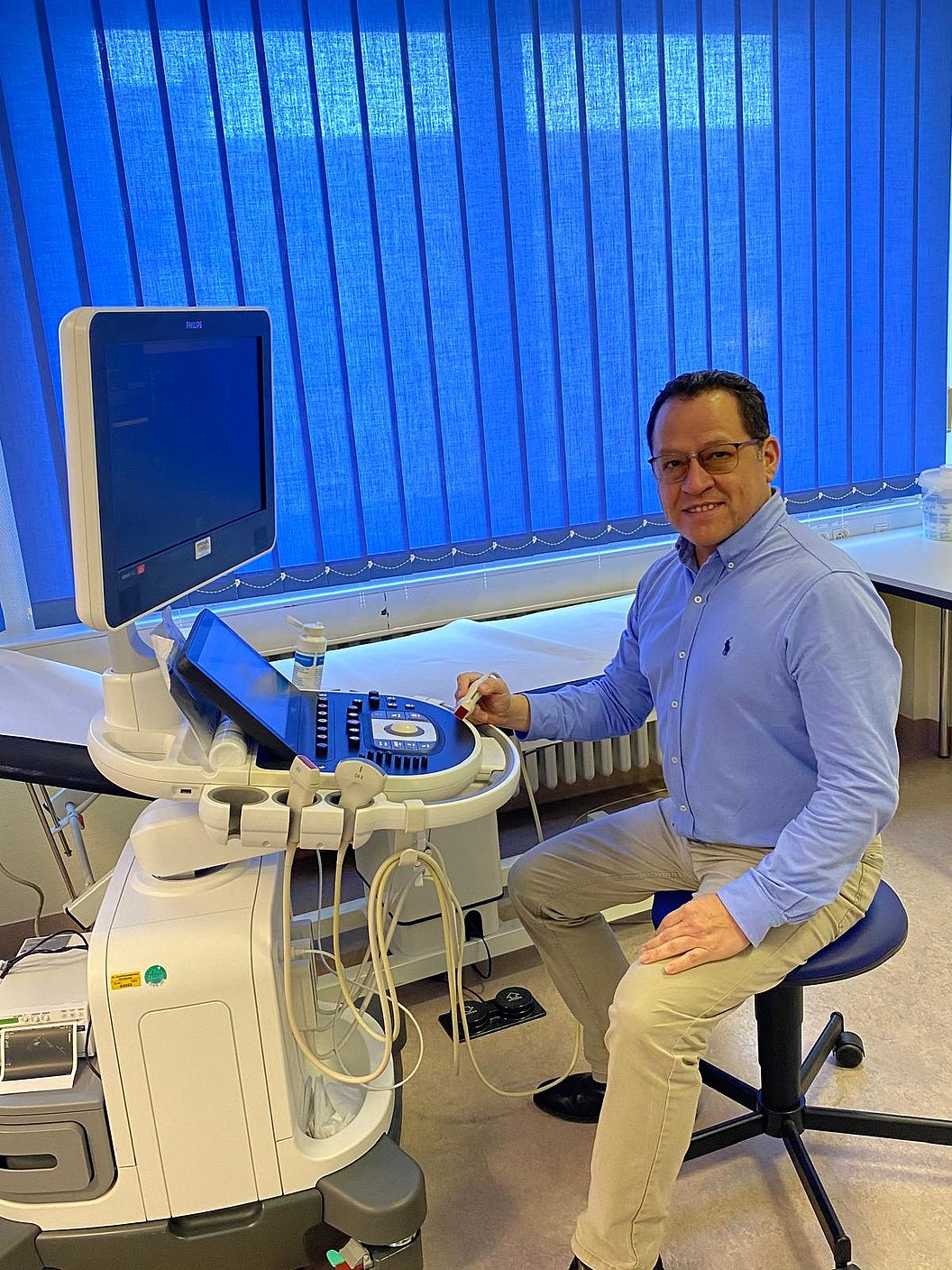 Ein Mann sitzt an einem Ultraschallgerät mit Monitor und Bedienelementen in einem Untersuchungsraum.