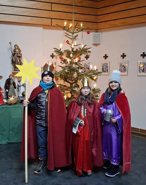 Drei Kinder in roten Umhängen und Kronen stehen vor einem geschmückten Weihnachtsbaum, eines hält einen großen gelben Stern an einem Stab.