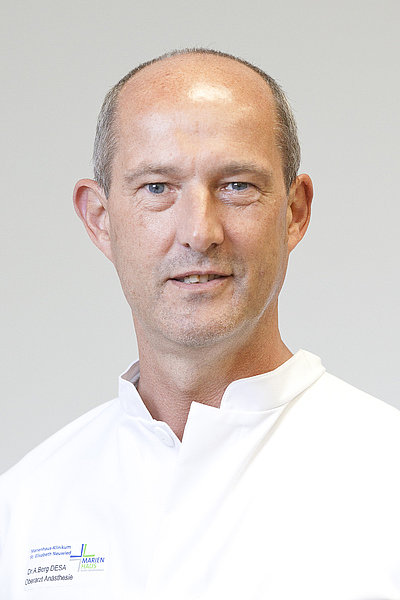 Rainer Kamprich trägt einen weißen medizinischen Kittel mit Namensschild und steht vor hellem Hintergrund