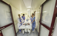Krankenhausflur mit drei medizinischen Fachkräften in blauer Arbeitskleidung, die eine liegende Person auf einer Trage schieben