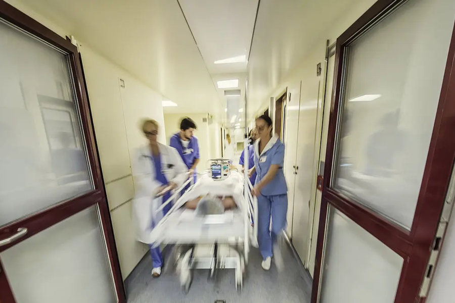 Krankenhausflur mit drei medizinischen Fachkräften in blauer Arbeitskleidung, die eine liegende Person auf einer Trage schieben