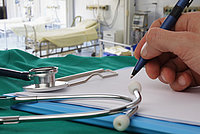 Hand mit Stift schreibt auf einem Klemmbrett neben einem Stethoskop auf einem grünen Tuch in einem Krankenhauszimmer