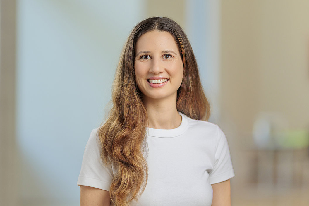 Frau mit langen, gewellten Haaren trägt ein weißes T-Shirt und steht vor einem unscharfen Hintergrund