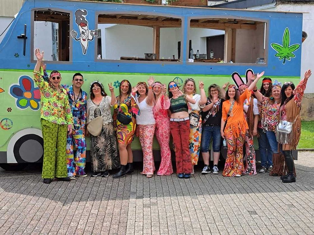 Gruppe von Menschen in bunter, gemusterter Kleidung vor einem blau-grünen Bus mit Blumen- und Hanfblatt-Malerei, die Hände erhoben.
