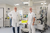 Zwei Personen in medizinischer Schutzkleidung stehen in einem hellen Behandlungsraum mit medizinischen Geräten und Schränken