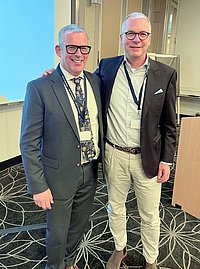Zwei Männer stehen nebeneinander in einem Konferenzraum. Der Mann links trägt einen dunklen Anzug mit einer gemusterten Krawatte, der Mann rechts trägt eine hellere Jacke und helle Hosen.