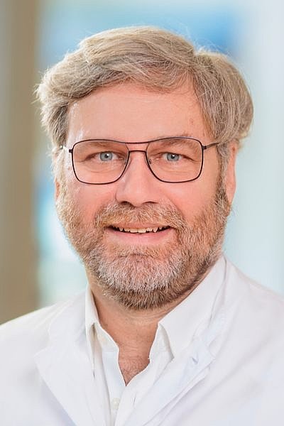 Dr. Walter Richter mit grauem Haar und Bart trägt weißen Arztkittel vor unscharfem Hintergrund.