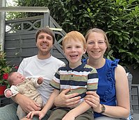 Eine Person hält ein Baby in einem weißen Anzug, ein Junge mit rotem Haar trägt ein gestreiftes T-Shirt, eine Frau in einem blauen Oberteil sitzt neben ihm. Im Hintergrund sind Pflanzen zu sehen.