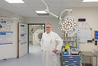 Ein Arzt in einem weißen Kittel steht mit den Händen in der Hüfte in einem Operationssaal. Im Hintergrund sind medizinische Geräte und ein CT-Scanner sichtbar.