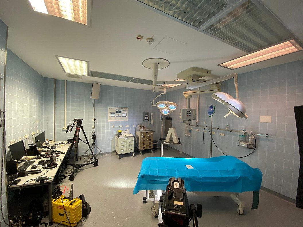 Operationssaal mit OP-Tisch, blau bedeckt, mehreren Lampen an der Decke, medizinischen Geräten und einem Tisch mit Computern und Kameras an der Wand