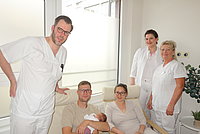 Drei in weißer Arbeitskleidung stehende Personen und ein sitzendes Paar, das ein Neugeborenes hält, in einem hellen Raum mit Fenster und Pflanze