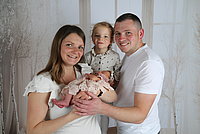 Eine Familie steht zusammen, die Mutter hält ein Baby in einem rosa Kleid, der Vater steht daneben. Ein Kind mit blonden Haaren steht zwischen ihnen.