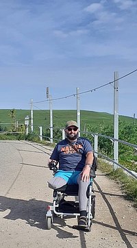 Mann mit Sonnenbrille und Basecap sitzt in einem Rollstuhl auf einem Weg mit grünem Hügel im Hintergrund