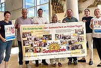 Sechs Personen stehen in einer Reihe und halten ein großes Banner mit dem Text 'Ein Kinderlachen - der Sonnenschein an trüben Tagen' und Bildern von Kindern. Das Banner feiert 20 Jahre Sonnenschein.