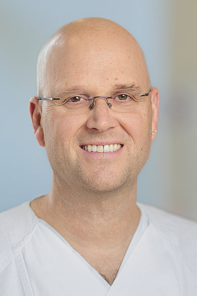Porträt eines glatzköpfigen Mannes mit Ohrring und weißem V-Ausschnittshirt vor hellem Hintergrund